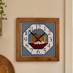 May include: A square wall clock with a wooden frame and a blue and white polka dot background. The clock face features a central illustration of a brown hen sitting on a nest of white eggs, with black numerals and black hour and minute hands.