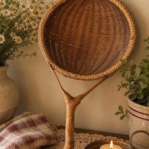 May include: A round, woven wicker fan with a natural wood branch handle. The fan features a tightly woven circular center with a braided border, attached to a sturdy, textured, Y-shaped wooden branch that serves as a handle for this decorative object.