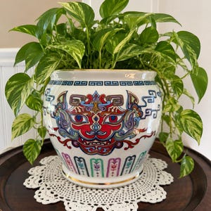 May include: A white ceramic planter featuring a colourful, symmetrical dragon-like face design in red, blue, and purple, with a gold-trimmed rim and base. The planter contains a lush, variegated green and yellow pothos plant with trailing vines.