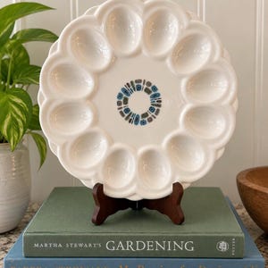 May include: A round white ceramic deviled egg platter with a scalloped edge and twelve oval indentations. The center of the plate features a circular pattern of small blue, teal, and brown abstract shapes arranged in a ring on the smooth surface.