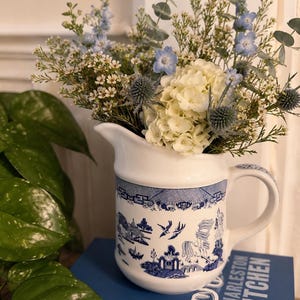 May include: A white ceramic pitcher featuring a blue willow pattern with birds and pagodas, filled with a bouquet of white hydrangeas, blue delphiniums, and eucalyptus. The pitcher rests on a blue book titled Charleston Kitchen.