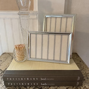 May include: Two silver-toned rectangular picture frames with beaded edges and beige fabric inserts featuring vertical grey stripes. The frames are displayed alongside a clear glass candle holder with a lit candle and a small glass jar of wooden matches.