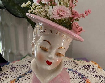 Peut inclure: Un vase en céramique en forme de tête représentant une personne portant un chapeau rose à larges bords orné d'une rose sculptée. Le visage présente des yeux fermés aux cils dorés, des lèvres rouges et un col rose. Le chapeau contient des fleurs.
