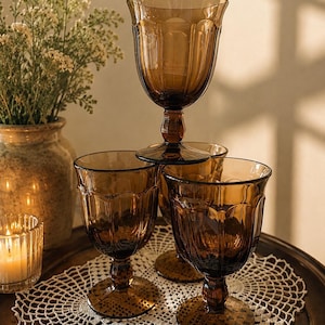 May include: A set of four amber-colored glass goblets with a scalloped rim design and a faceted stem. The glasses feature a translucent brown hue and a textured base, arranged in a stacked formation to display the vertical paneling on the bowls.