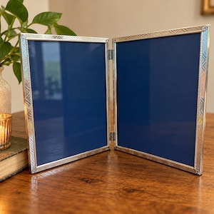 May include: A double-hinged silver-toned metal picture frame featuring a textured geometric border pattern. The frame consists of two rectangular panels connected by a central hinge, each containing a solid dark blue insert behind clear glass.