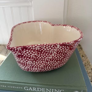 May include: A heart-shaped ceramic bowl featuring a cream-colored interior and a deep red exterior pattern consisting of small, dense white dots. The bowl has a smooth, curved rim and a deep basin, showcasing a consistent dotted texture throughout.