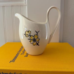 Könnte beinhalten: Ein cremefarbenes Milchkännchen aus Keramik mit einem zarten, handgemalten Blumenmuster in Gelb und Schwarz. Das Kännchen hat einen geschwungenen Henkel und einen spitzen Ausguss sowie eine glatte, glänzende Oberfläche mit feiner Craquelé-Struktur.