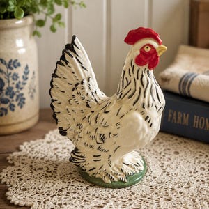 Puede incluir: Una figura de gallina de cerámica con cuerpo blanco y detalles de plumas negras, cresta y barbillas de color rojo brillante y pico amarillo. El ave se apoya sobre una pequeña base verde, mostrando un plumaje texturizado con plumas negras en la cola.