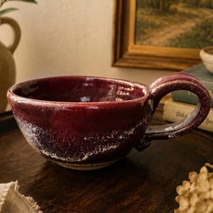 May include: A deep burgundy ceramic mug with a glossy finish and a thick, rounded handle. The exterior features a textured, mottled white glaze effect near the base, contrasting with the dark red upper portion of the bowl-shaped vessel.