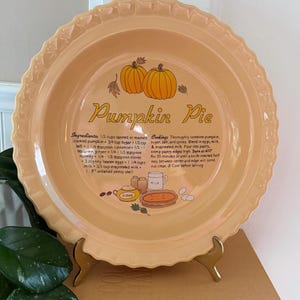 May include: A light orange ceramic pie dish with a fluted rim. The centre features a graphic of two pumpkins, autumn leaves, and a recipe for Pumpkin Pie, including ingredients like 1/2 cup sugar and 1/2 teaspoon ginger, plus cooking instructions.
