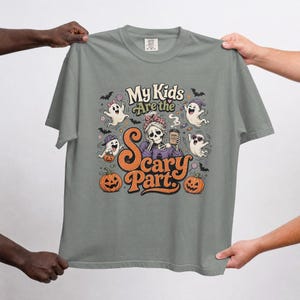 May include: A sage green t-shirt featuring a graphic print with the text My Kids Are the Scary Part. The design includes a skeleton character holding a coffee cup labeled Mom Fuel, surrounded by four cartoon ghosts and two orange pumpkins.