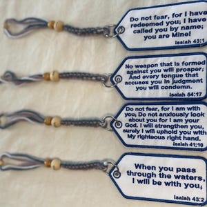 May include: Four white fabric tags with blue embroidered borders and text. Each tag features a different Bible verse: Isaiah 43:1, 54:17, 41:10, and 43:2. The tags are attached to braided grey and white cord loops with small wooden beads.