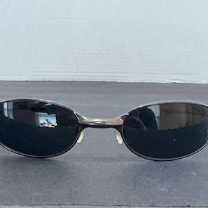 Puede incluir: Gafas de sol de forma ovalada con una montura de metal gris plomo y lentes oscuras y reflectantes. El diseño incluye un puente fino que conecta ambas lentes y almohadillas nasales integradas, ofreciendo un aspecto elegante y pulido.