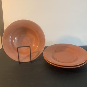 May include: A set of three terracotta-coloured ceramic bowls with a smooth, glossy finish. One bowl is displayed upright on a black metal wire stand, while the other two bowls are stacked horizontally next to it, showcasing their wide, circular rims.