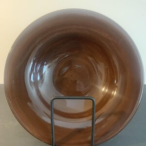 May include: A deep, circular brown bowl with a smooth, glossy finish. The bowl features a wide, curved rim and a concave centre. It is supported by a simple, black metal wire stand that holds the bowl in an upright, vertical display position.