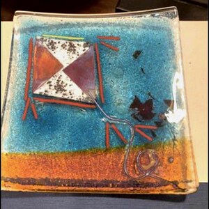May include: A square glass dish featuring a vibrant blue and orange background. The center displays a kite graphic with white and maroon triangular sections, accented by red lines and a trailing string that curls toward the bottom right corner.