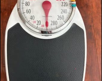 May include: A white and black analog bathroom scale with a round dial face featuring the brand name Taylor. The dial displays numbers from 0 to 330 lbs in increments of 20, with a red needle pointing to zero and two small colored markers on the side.