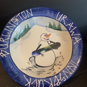 Puede incluir: Un cuenco de cerámica redondo con borde azul oscuro con pintura blanca salpicada y el texto BURLINGTON, URAWA e INNSBRUCK. El centro muestra un muñeco de nieve pintado a mano sosteniendo una tabla de snowboard Burton en una colina.