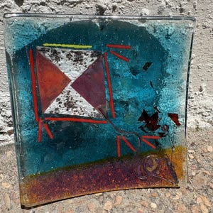 May include: A square fused glass art piece featuring a blue background with a central kite design. The kite is composed of white and dark red triangular segments with black speckles. A red string trails down to a small dark red floral element.