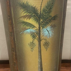 Peut inclure: Une corbeille à papier haute et effilée avec une surface texturée jaune doré peinte d'une illustration centrale de palmier vert. Le récipient possède un bord tressé marron foncé, des supports d'angle verticaux en bois et trois petits pieds.