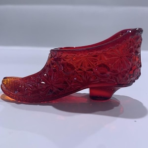 May include: A decorative shoe figurine crafted from translucent ruby red glass. The exterior surface features a raised, repeating geometric starburst and circular pattern across the entire body, including a small, integrated block heel.