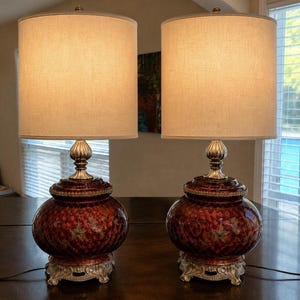 May include: A pair of table lamps featuring cylindrical beige fabric shades and bulbous, deep red ceramic bases with intricate gold-toned floral patterns. The lamps stand on ornate, silver-colored metal bases with a matching silver neck detail.