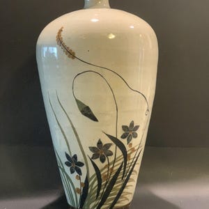 May include: A tall, off-white ceramic vase with a narrow neck and a flared rim. The body features a hand-painted botanical design with dark blue flowers, green grass-like blades, and thin brown stems with small circular accents on a smooth surface.