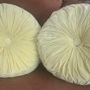 May include: Two round, pale yellow cushions featuring a pleated design that radiates from a central fabric-covered button. The surface of each cushion has a soft, velvet-like texture with deep folds creating a circular, tufted pattern across the front.