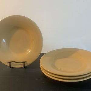 May include: A set of four round, solid light tan ceramic bowls with smooth, curved edges and a glossy finish. One bowl is displayed upright on a black metal stand, while the other three are stacked neatly in a pile to the right side.
