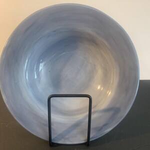 May include: A round ceramic bowl featuring a marbled pattern in shades of light and medium blue. The bowl is displayed upright, resting on a thin, black metal wire stand that supports the curved base and provides a stable vertical orientation.