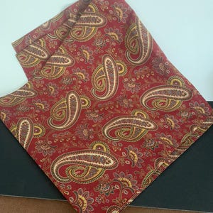 May include: A square fabric cloth featuring a deep red background with a repeating paisley pattern in shades of gold, cream, and dark blue. The intricate design includes swirling teardrop shapes and floral motifs distributed across the surface.