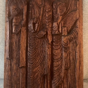 Pode incluir: Uma placa de parede retangular de madeira com um relevo de três figuras em pé vestidas com longas túnicas drapeadas. A madeira castanho-escura apresenta um padrão de grão vertical proeminente, com as figuras dispostas lado a lado.