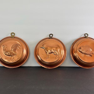 May include: Three round copper molds featuring embossed animal designs. Each mold has a small brass hanging loop at the top. The left mold depicts a rooster, the center mold shows a cow, and the right mold features a fish in relief.