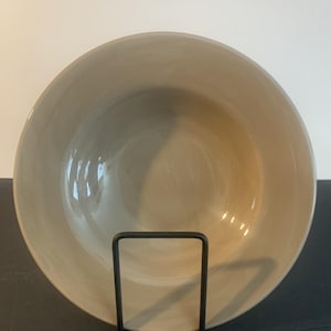 May include: A round, smooth, light beige ceramic bowl with a wide rim and a glossy finish. The bowl is held upright by a simple black metal display stand, showcasing its clean, circular shape and neutral colour palette for a clear view.