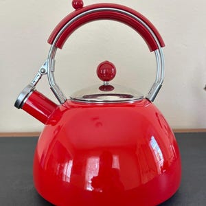 May include: A bright red stovetop kettle with a glossy finish, featuring a chrome-plated lid with a red spherical knob, a matching red handle with a chrome frame, and a spout with a hinged mechanism for opening and closing the kettle opening.
