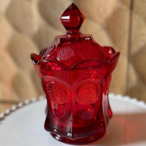 Puede incluir: Un tarro de cristal rojo vibrante con tapa a juego, que presenta un borde festoneado y medallones circulares en relieve con emblemas de águila. El recipiente translúcido tiene un cuerpo facetado y un remate puntiagudo en la tapa.