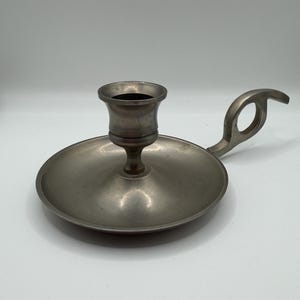 May include: A silver-toned metal chamberstick candle holder featuring a wide, circular drip tray base, a central cylindrical candle socket, and a curved handle with a circular cutout for a secure grip, all finished in a smooth, matte metallic texture.