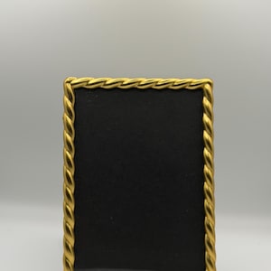 May include: A rectangular picture frame with a metallic gold finish and a decorative twisted rope pattern along the border. The frame features a solid black insert, providing a stark contrast to the bright, reflective gold edges of the frame.