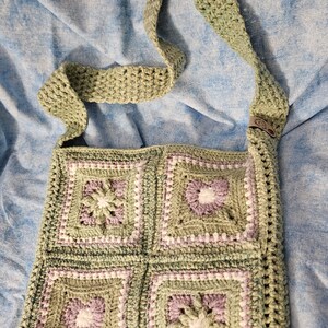 May include: A handmade crochet crossbody bag featuring four square motifs in sage green, lavender, and white. The bag has a long shoulder strap and a small leather-like tag on the side. The squares display alternating floral and heart patterns.