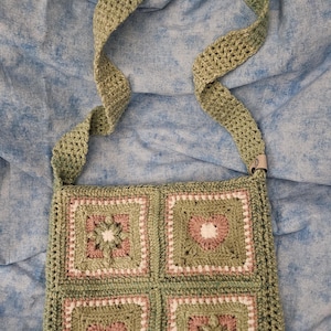 May include: A square crochet bag featuring four granny squares in sage green, dusty pink, and white. The squares display alternating floral and heart motifs. The bag includes a wide, matching sage green crochet shoulder strap and a small tag.