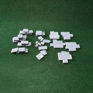 Op de afbeelding: Een verzameling kleine, lichtgrijze, vierkante en rechthoekige tegels gerangschikt in verschillende clusters. Deze individuele stukken hebben een gladde, licht glanzende afwerking en zijn gegroepeerd in verschillende formaties die doen denken aan miniatuur plaveiselstenen.