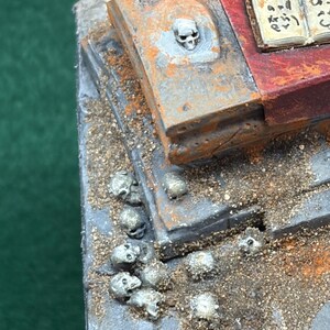 Może przedstawiać: Miniaturowa diorama przedstawiająca zniszczony kamienny ołtarz z czerwoną powierzchnią, otwartą księgę z nieczytelnym tekstem oraz dwie małe świece. Pojedyncza czaszka jest wbudowana w ołtarz, a stos małych czaszek spoczywa na podłożu.