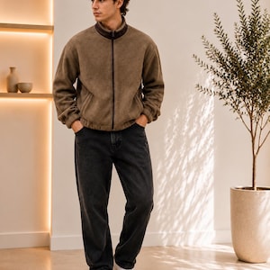 May include: A brown fleece zip-up jacket with a high collar and dark brown trim, paired with charcoal gray denim jeans and white sneakers. The jacket features a textured material, a full-length front zipper, and long sleeves with ribbed cuffs.