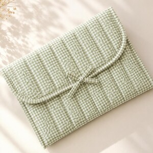 Puede incluir: Un estuche acolchado rectangular con un patrón de cuadros vichy en color verde salvia claro y blanco. El estuche tiene una solapa redondeada que se asegura con un lazo de tela a juego. El exterior presenta costuras verticales en toda la superficie.