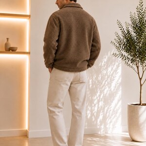 May include: A brown long-sleeved fleece jacket with a collar and a full-length front zipper, paired with off-white trousers and plain white sneakers. The jacket features a textured, fuzzy material and is worn over the light-colored pants.