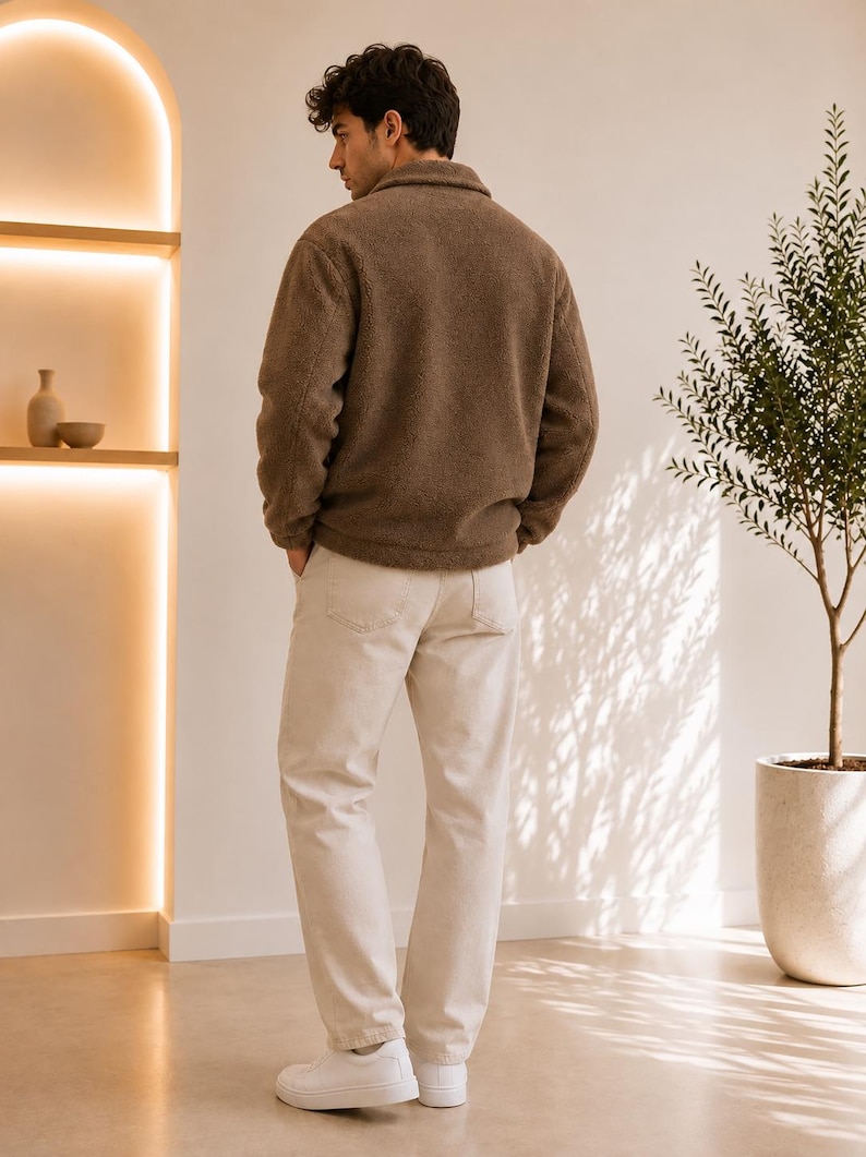 May include: A brown long-sleeved fleece jacket with a collar and a full-length front zipper, paired with off-white trousers and plain white sneakers. The jacket features a textured, fuzzy material and is worn over the light-colored pants.