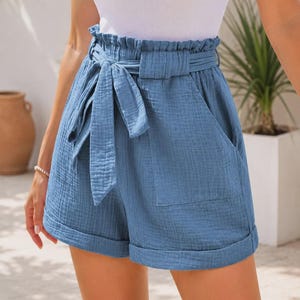 Puede incluir: Pantalones cortos azules de talle alto con cintura tipo paper bag, cinturón de tela a juego, dos bolsillos laterales y dobladillos con vuelta. Los pantalones cortos están hechos de una tela texturizada y se combinan con una camiseta blanca sin mangas.