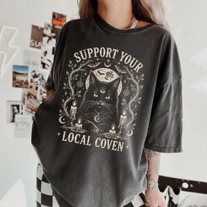 Könnte beinhalten: Ein anthrazitfarbenes Kurzarm-T-Shirt mit einem Grafikdruck einer dreiaugigen Katze, umgeben von Kerzen, Bäumen und einem Mond. Der Text "SUPPORT YOUR LOCAL COVEN" ist in einer verwaschenen cremefarbenen Schrift über und unter der Katze gedruckt.