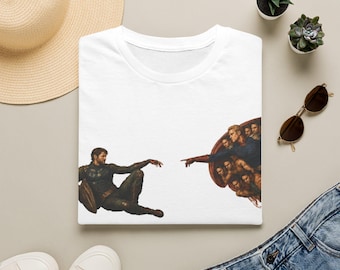 May include: A white t-shirt featuring a graphic print inspired by The Creation of Adam, depicting a dark-clad figure reaching toward a group of figures in blue and red. The design is centered on the chest of the short-sleeved cotton garment.