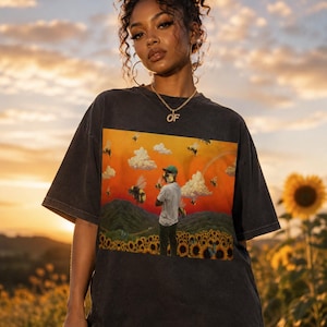 Pode incluir: Uma t-shirt cinzento-carvão de manga curta com uma impressão gráfica retangular de uma pessoa num campo de girassóis sob um céu laranja ao pôr do sol, cheio de abelhas a voar e nuvens brancas. A camisola tem um decote redondo.